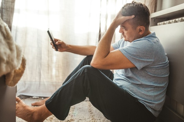 Stressed man sits on the floor looking at his phone.