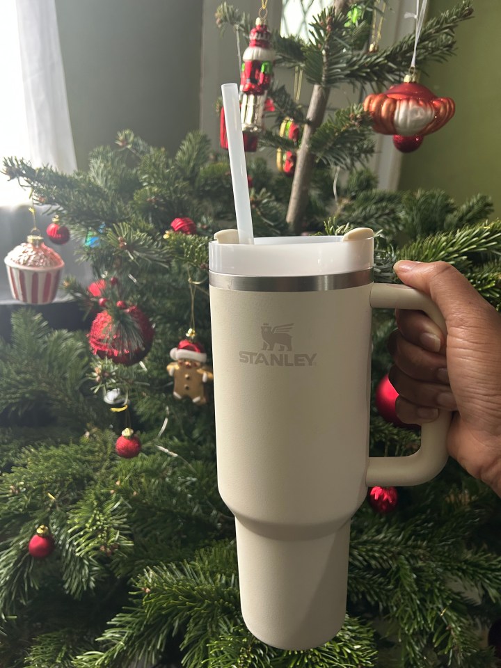 A hand holding a Stanley travel mug with a straw in front of a decorated Christmas tree.