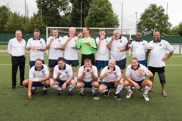 Group of men in England football jerseys eating and drinking on a soccer field.