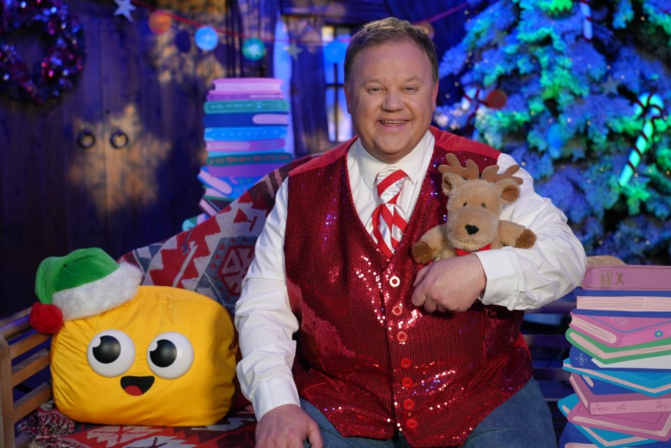 A man in a red sequined vest holds a reindeer plushie, sitting on a couch with stacks of books and a yellow character pillow.