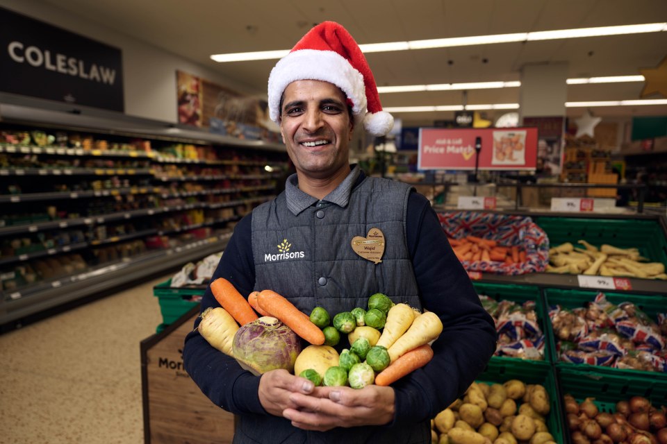 A Morrisons employee wearing a Santa hat holds a selection of Christmas vegetables.