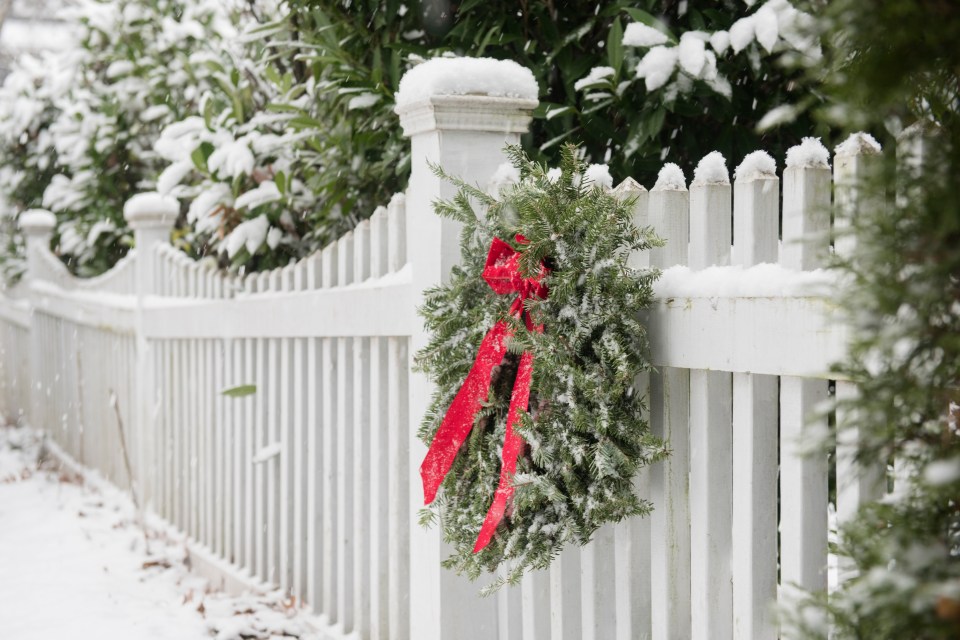 Snow-covered evergreen wreath with a red ribbon on a white fence.