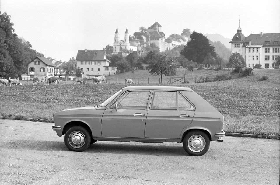 Black and white photo of a Peugeot 104 car parked in a field with cows and a castle in the background.