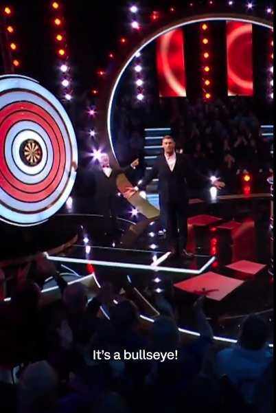 Two men on a game show stage celebrating a bullseye on a large dartboard.