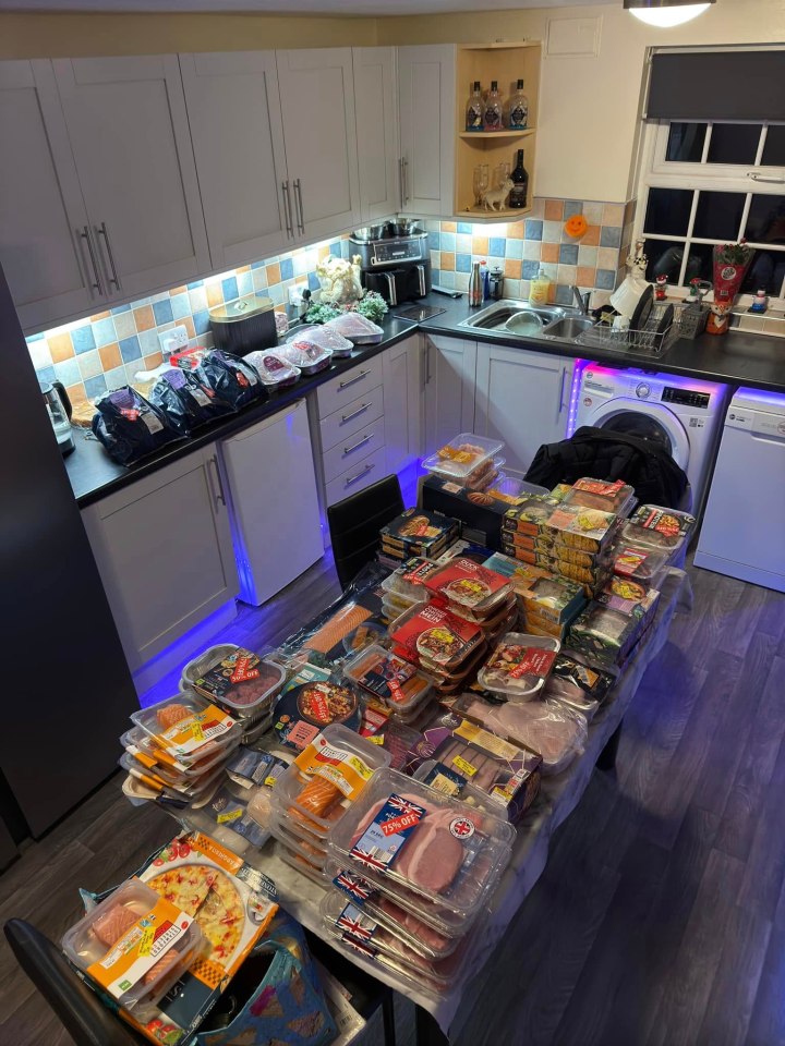 A kitchen table piled high with groceries, including meat, fish, and prepared meals.