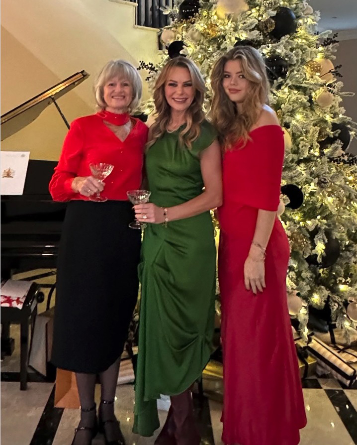 Three women in holiday dresses pose by a Christmas tree.