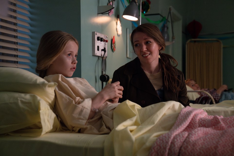 A young girl in a hospital gown lies in a bed, holding hands with a woman.