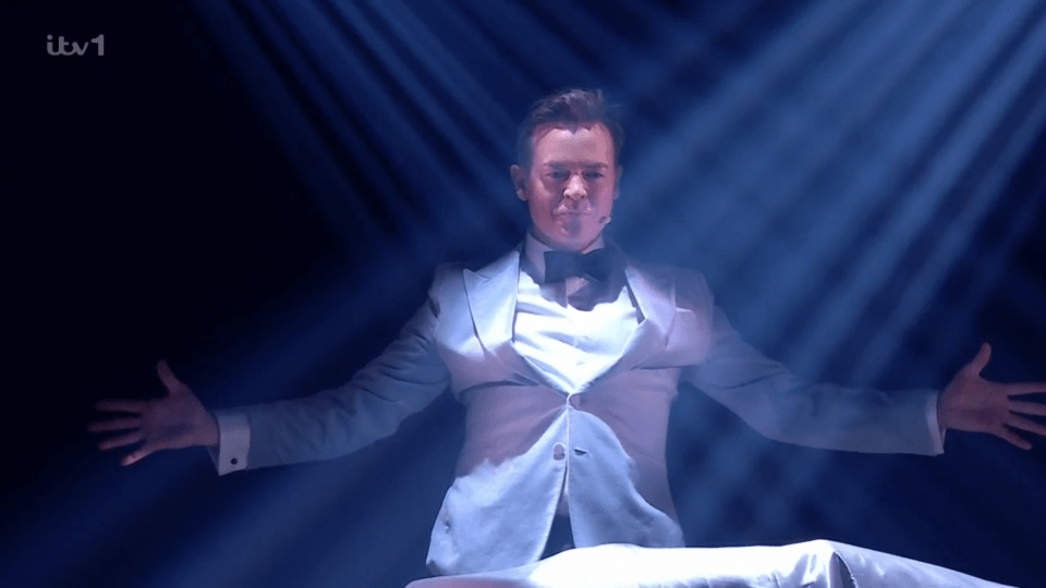 A man in a white tuxedo performs a magic trick on stage.