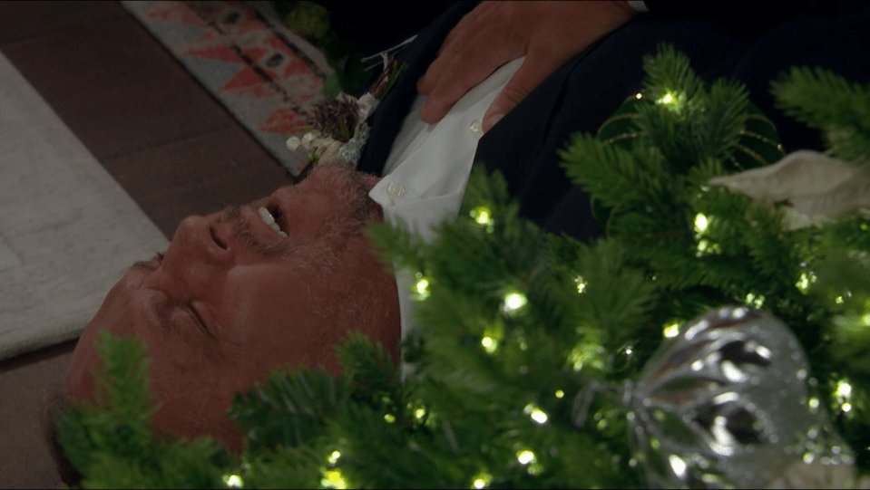 Man in suit lying on the floor near a Christmas tree, appearing to be unconscious.