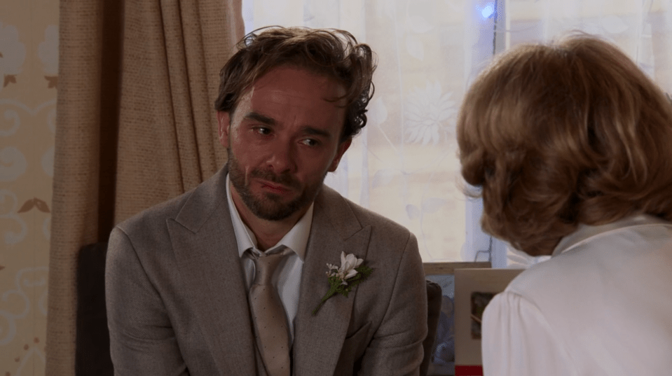 A man in a suit, wearing a boutonniere, sits and speaks with a woman.