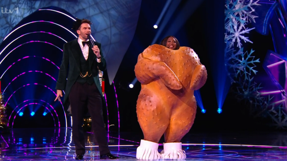 A person in a roasted chicken costume on a game show stage with a host.