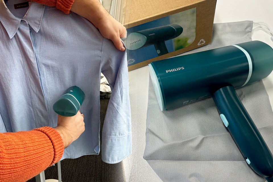 A person holding a handheld steamer, looking at the garment hanging in the background.