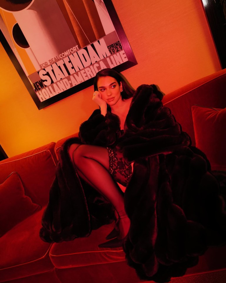 Dua Lipa in a fur coat on a red couch.