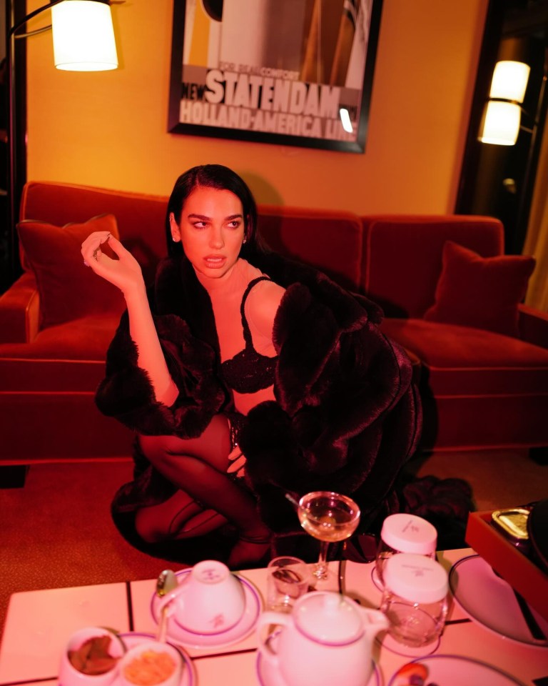 Dua Lipa in a fur coat, sitting on a red couch.