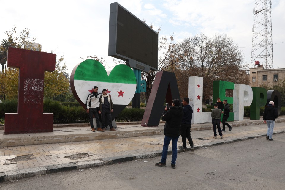 Locals in Aleppo stand in front of a sign painted with the colours of HTS' flag