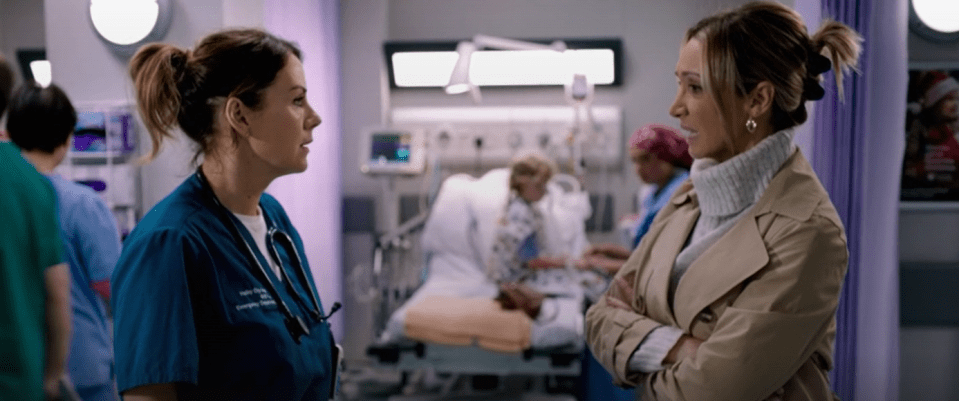 Two women in a hospital, one in scrubs, the other in a coat, having a serious conversation.