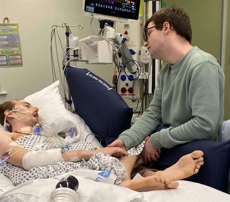 A young man in a hospital bed, holding his partner's hand.