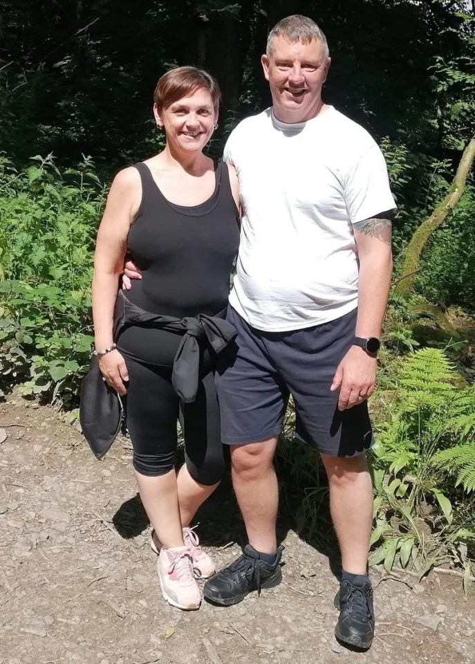 A woman and a man stand together on a path in a wooded area.