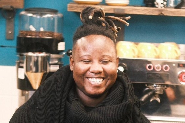 A smiling person with dreadlocks sits at a cafe counter with a small cup of coffee.