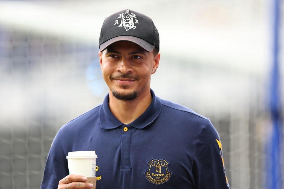 Dele Alli, wearing an Everton polo shirt and baseball cap, smiles while holding a coffee cup.