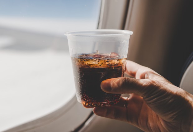 Hand holding a plastic cup of cola with ice by an airplane window.