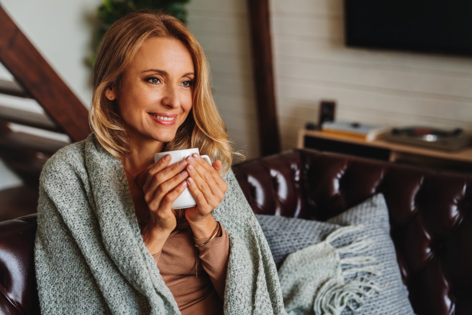 Close up of dreaming middle aged woman sitting in living room with cup of coffee or tea enjoying under blanket