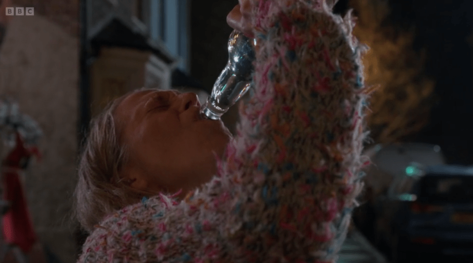 A woman in a colorful sweater drinks from a bottle outdoors at night.