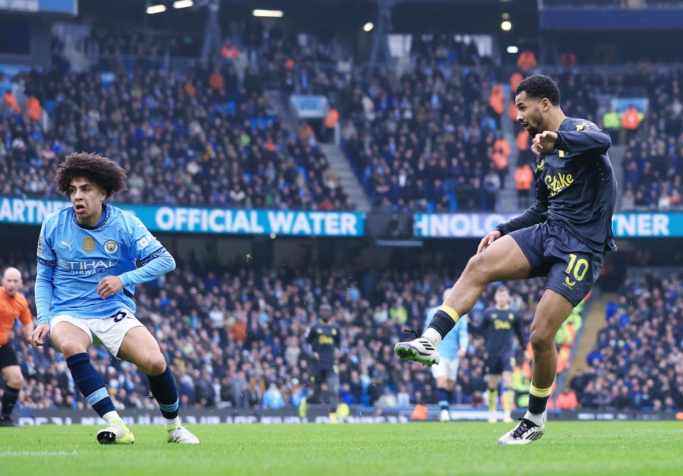 A soccer player in a dark jersey kicks the ball while another player in a light blue jersey defends.