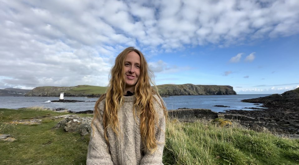 Woman with long blonde hair smiles in front of a coastal landscape.