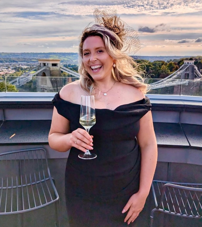 Woman in black dress holding champagne, smiling on rooftop with Clifton Suspension Bridge in background.