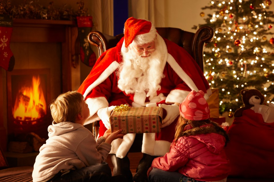 Santa Claus giving presents to two children by a fireplace.