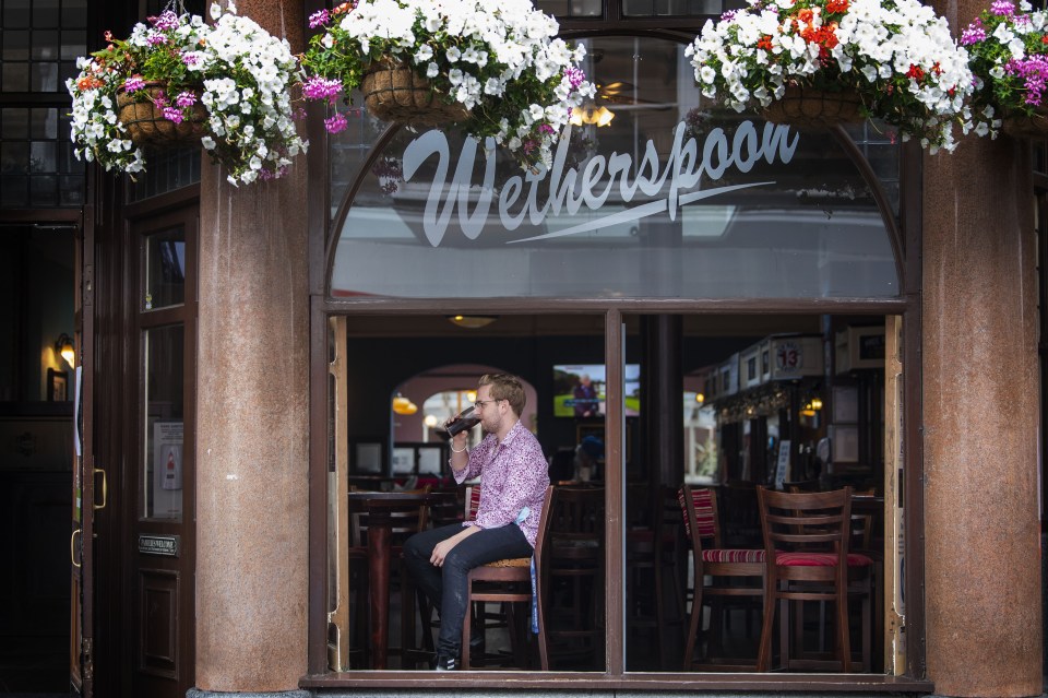 A man sits at a table inside a Wetherspoon pub, drinking from a pint glass.