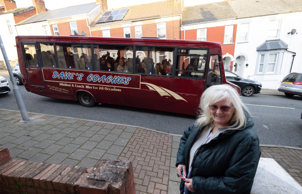 A woman stands near a tour bus in front of a house.