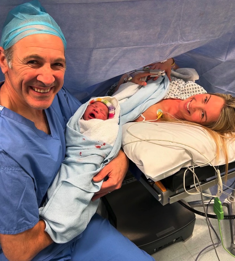 A man holds a newborn baby while a woman lies in a hospital bed.