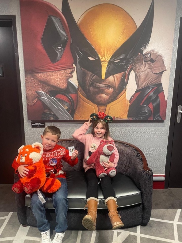 Two children sit on a couch in front of a mural of Deadpool, Wolverine, and another character.