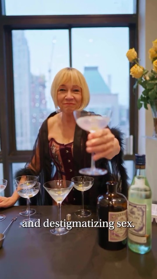 A woman holds up a martini glass, several other martini glasses are on a table in front of her.