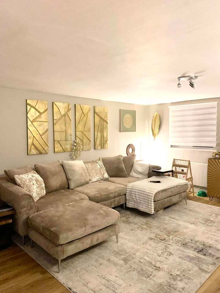 A taupe sectional sofa with a matching ottoman sits on a large area rug in a living room.  Gold abstract art hangs on the wall above the sofa.