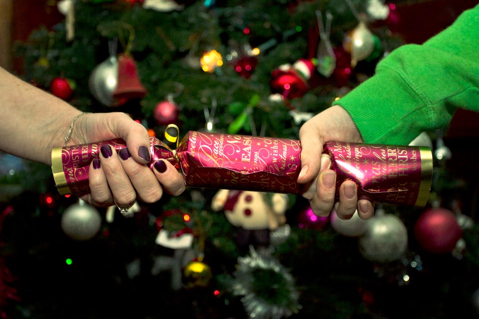 Two hands pulling a Christmas cracker in front of a decorated Christmas tree.