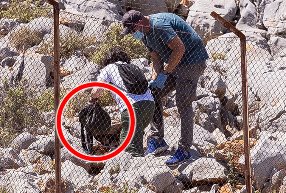 Two people retrieving a bag from behind a chain-link fence.