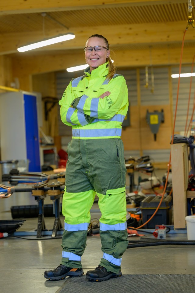 Woman in safety gear smiling with arms crossed in a workshop.