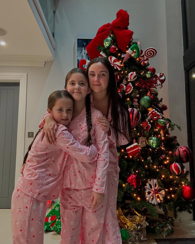 Woman and two children in matching pink Christmas pajamas by a decorated Christmas tree.