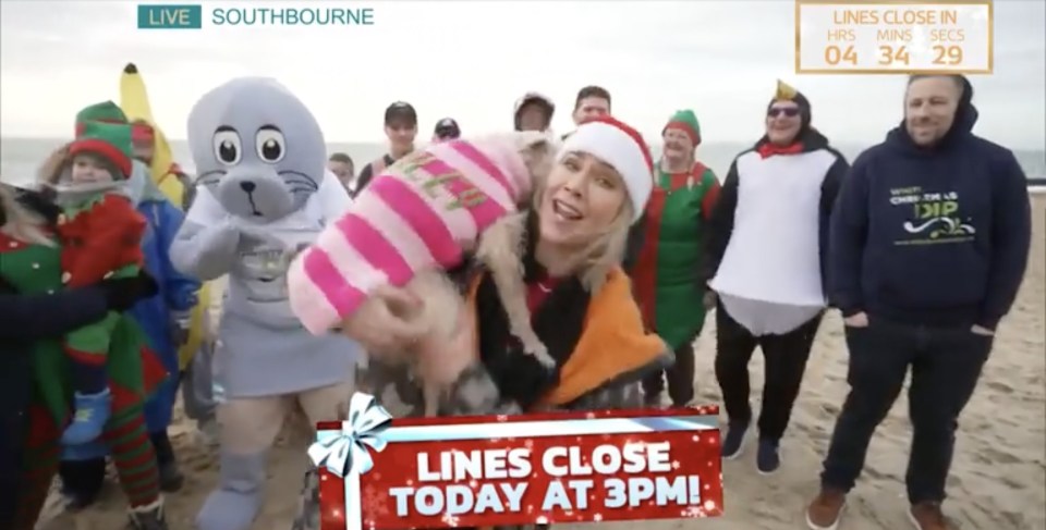 A woman holds a dog wearing a pink and white sweater on a beach with people in Christmas costumes.