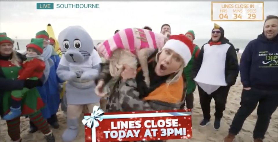 A woman in a Santa hat holds a dog on a live TV show on a beach, surrounded by people in Christmas costumes.