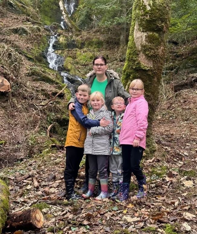 A woman and her four children stand together in a wooded area near a waterfall.