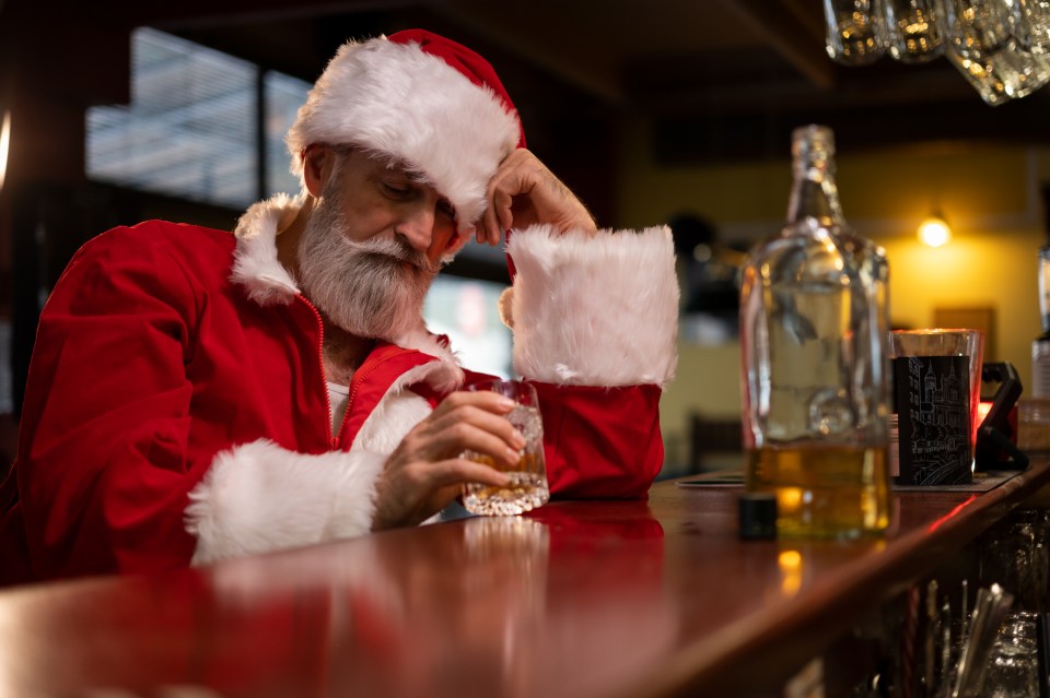 A man dressed as Santa Claus sits at a bar, looking dejected and holding a glass of whiskey.