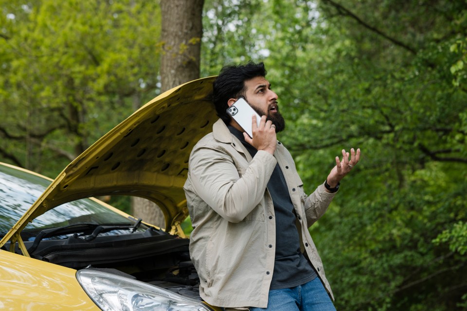 Man on roadside phone call, car hood up.