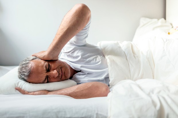 A man lies in bed with his hands on his head, appearing to have a headache.