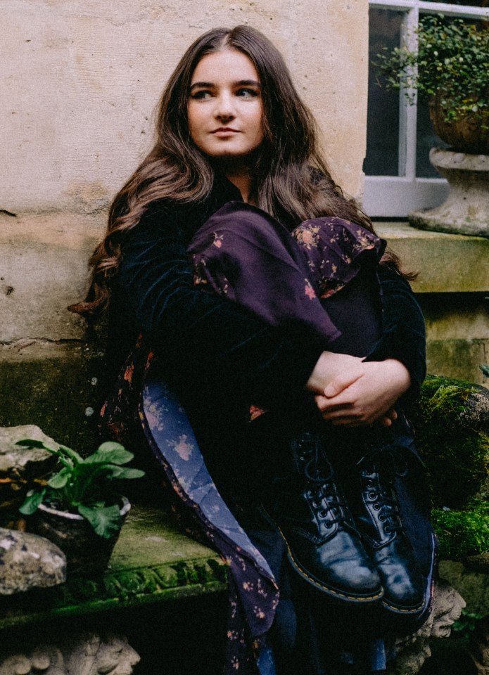 A young woman with long brown hair sits outdoors, wearing a dark floral dress and Doc Martens.