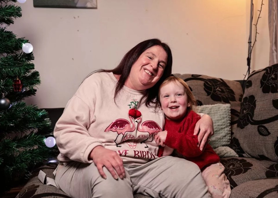 A mother and her young daughter sit together on a couch in front of a Christmas tree.