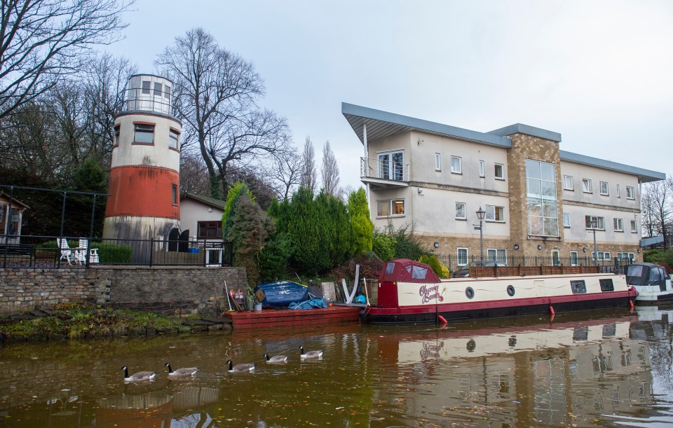 A canal-side lighthouse next to a building and narrowboat, with geese swimming in the water.